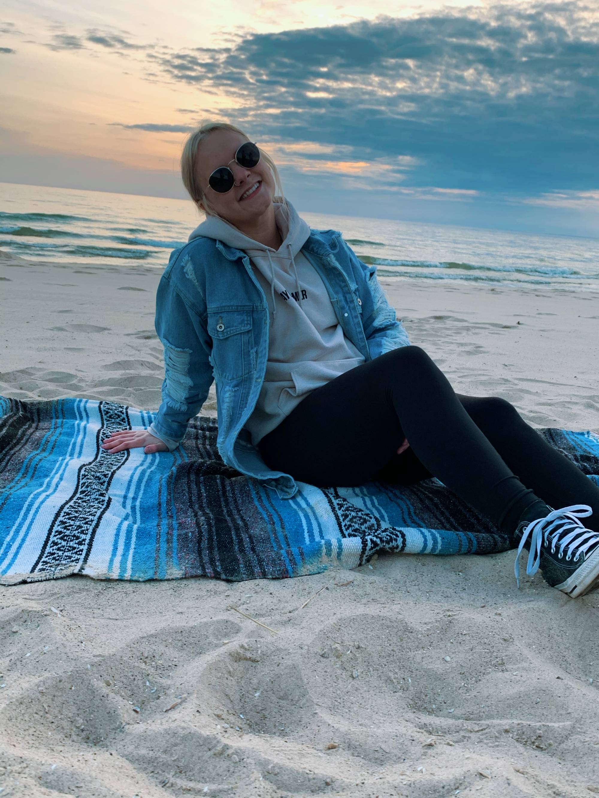 Girl sitting on the beach on a blanket wearing sunglasses, a denim jacket and black pants. There is a sunset in the background.